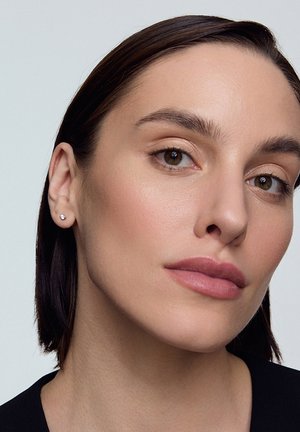 Metalowe kolczyki w kształcie okręgu, noszone na lewym uchu, o polerowanym wykończeniu. Osoba ma średniej długości ciemne włosy i neutralny makijaż.