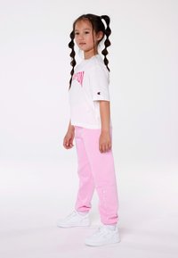 Camiseta blanca con logo "Champion" en rosa, combinada con pantalones de chándal rosa y zapatillas blancas. Corte relajado y texturas de tela suaves.