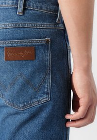 Vaqueros de mezclilla azul oscuro con un parche de cuero marrón de Wrangler en el bolsillo trasero, con costuras amarillas y un diseño clásico de bolsillo.