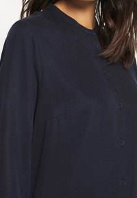 Donna che indossa una camicia blu navy con bottoni, maniche lunghe e collo senza risvolto, capelli parzialmente visibili sulla spalla.