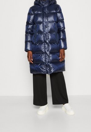 Osoba ubrana w długą, błyszczącą granatową kurtkę puchową z kapturem, czarne szerokie spodnie oraz białe sneakersy na platformie, stojąca na jednolitym tle.