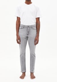 Jean gris à coupe slim avec une texture légèrement délavée, associé à un t-shirt blanc uni à manches courtes. Pieds nus.
