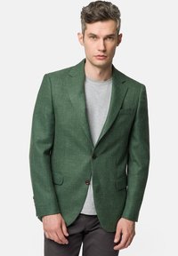 Zielony żakiet w wzór rybiej ości z klapami, dwoma guzikami i kieszenią na piersi. Noszony na jasnoszarym t-shircie, w zestawieniu z ciemnymi spodniami.