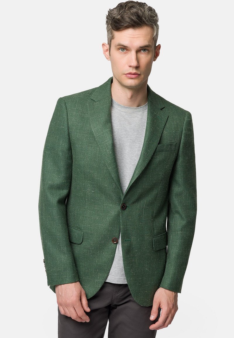 Zielony żakiet w wzór rybiej ości z klapami, dwoma guzikami i kieszenią na piersi. Noszony na jasnoszarym t-shircie, w zestawieniu z ciemnymi spodniami.