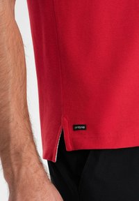 Rød polo t-shirt med tekstureret stof, korte ærmer, side splitter og et sort "ombre" mærke nær kanten. Sorte bukser delvist synlige.