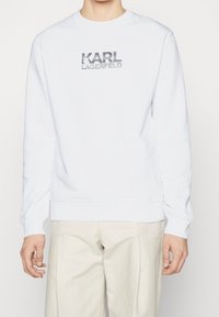 Weißer Sweatshirt aus weichem Stoff mit Rundhals. Mit silbernem Logo-Text "KARL LAGERFELD" auf der Vorderseite und gerippten Bündchen.