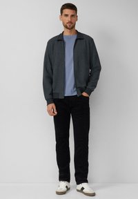Jacke mit Knopfleiste aus dunkelgrauem, strukturiertem Stoff, getragen über einem hellblauen Hemd. Schwarze Hose und weiße Sneakers mit schwarzen Akzenten vervollständigen den Look.
