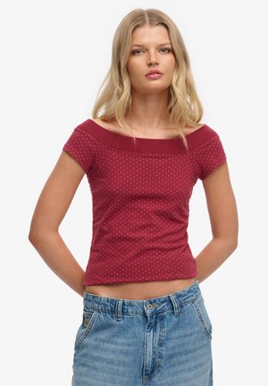 Jeune femme aux cheveux blonds longs portant un haut rouge ajusté à épaules dénudées avec des pois blancs et un jean bleu taille haute, debout les mains derrière le dos.