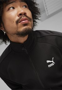 Svart zip-up jacka med en texturerad yta, med en vit Puma-logotyp på vänster bröst och en hög krage.