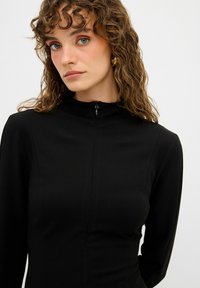 Schwarzes, figurbetontes Kleid mit hohem Kragen, langen Ärmeln und Reißverschlussdetails am Hals. Weicher Stoff, minimalistische Nähte. Das Model hat lockige Haare.