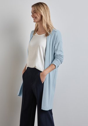 Cardigan ouvert bleu clair en tissu doux, avec des manches longues et une coupe décontractée, associé à un t-shirt blanc et un pantalon sombre.