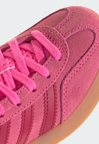 Gros plan sur une basket en suède rose avec des lacets assortis et une semelle en gomme, montrant des coutures détaillées et une matière supérieure texturée.