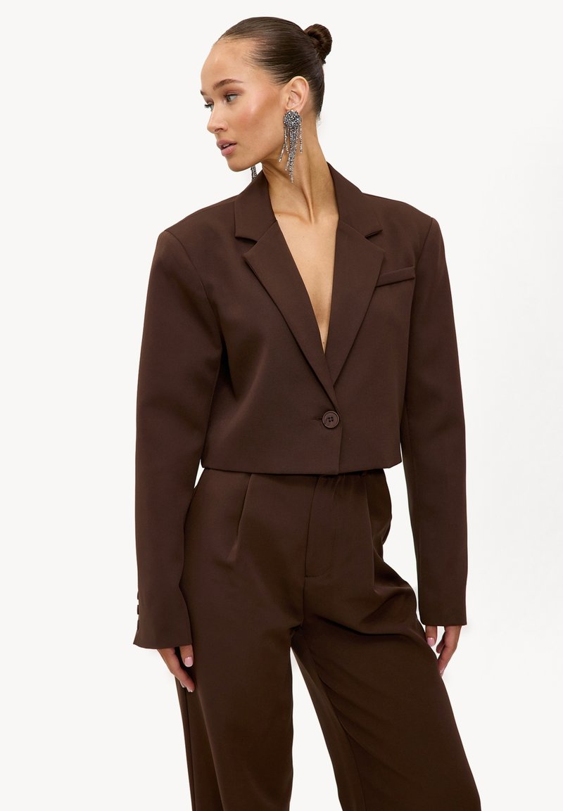 Femme avec une coiffure relevée portant un blazer court oversize marron foncé et un pantalon taille haute assorti, assortis de longues boucles d'oreilles pendantes en argent.