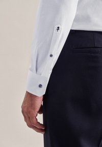 Męska ręka u boku ubrana w białą koszulę z drobną czarną różą wyszytą na mankiecie oraz ciemnogranatowe spodnie.