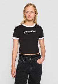 Juoda trumpa marškinė su balta kontrastine apykakle ir rankogaliais, su užrašu "Calvin Klein" ir "endless city" išspausdintu ryškiomis baltomis raidėmis.