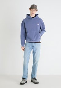 Blue hoodie with kangaroo pocket and ribbed cuffs, paired with light blue straight-leg jeans and grey shoes. Hat shows subtle logo detail.