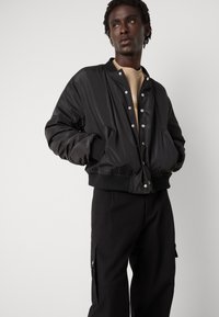 Black bomber jacket with a glossy finish, snap button closure, ribbed cuffs, and two side pockets, worn over a light beige shirt.