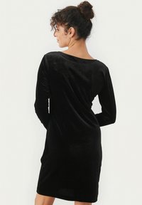 Robe en velours noir à manches longues et décolleté rond, présentant un design à dos drapé qui met en valeur sa texture lisse.