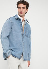 Camisa de denim azul clara com um fecho de botão à frente, um bolso no peito e acabamentos texturizados. Apresenta um colarinho clássico e um corte descontraído.