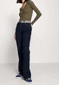 Dunkelblaue Jeans mit ausgestelltem Bein und hohem Bund, kombiniert mit einem figurbetonten, olivgrünen, gerippten Langarmshirt und einem gemusterten Gürtel. Blaue Turnschuhe runden das Outfit ab.