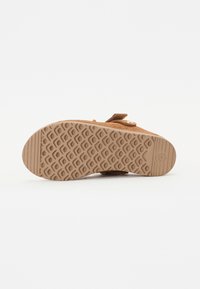 Beige semsket sandal med en teksturert gummisåle. Har en stropp og metallkomponenter, med et mønstrete dekk for grep.
