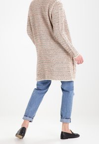Lång stickad cardigan i beige med ett strukturerat mönster, bärs över ljusblå jeans. Svarta slip-on skor fulländar outfiten.