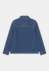 Blå denimjacka som ligger platt med baksidan upp, med långa ärmar, knäppta manschetter och en liten rektangulär broderad logga nära övre mitten.