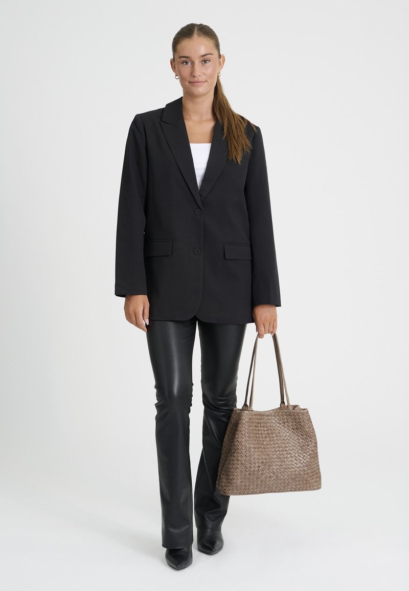 Jeune femme portant un blazer noir et un pantalon en cuir, tenant un grand sac cabas tressé marron, debout sur un fond blanc.
