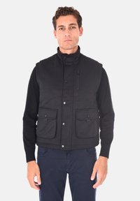 Gilet nero con tessuto testurizzato, colletto alto e tasche frontali. Tasca con zip sul lato sinistro del torace. Indossato sopra una camicia a maniche lunghe.