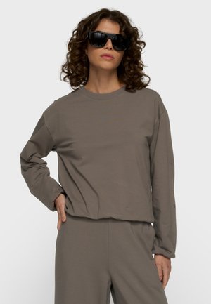Frau mit lockigem Haar, die übergroße schwarze Sonnenbrille und ein passendes, lockeres taupefarbenes Langarmoberteil und Hose trägt, steht mit einer Hand in der Hüfte.
