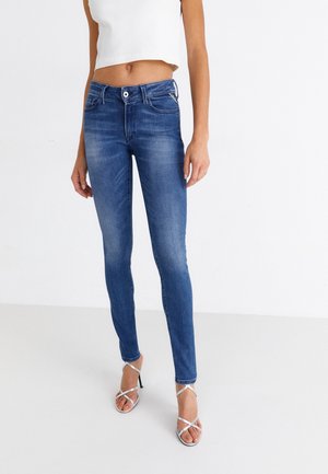 Vrouw die een spijkerbroek met hoge taille in blauw draagt, met vervaagde details, gecombineerd met witte sandalen met hoge hak en bandjes, en een cropped witte mouwloze top.