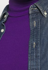 Denim jacket with a faded blue color, featuring a collar and visible stitching. Underneath, a ribbed purple turtleneck is visible.