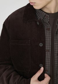 Veste en velours côtelé marron avec un col, dotée d'une poche poitrine et de boutons noirs, superposée sur une chemise à carreaux à boutons dans des tons sombres.