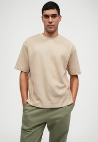 Beige cotton t-shirt with short sleeves and a round neckline. Ribbed texture, relaxed fit, worn with green pants.