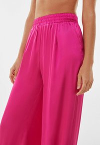 Flowy pink satin pants with an elastic waistband, featuring side pockets and a smooth texture, partially shown from the waist down.