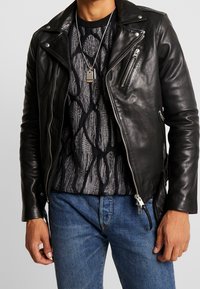 Svart läderbikerjacka med silverfärgade dragkedjor, lagd över en texturerad grå tröja och blå denimjeans. Fäst hundtag-halsband.