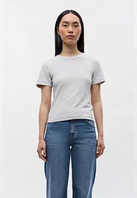 Giovane donna con capelli neri lisci indossa una semplice maglietta grigia chiara a maniche corte e jeans in denim blu, in piedi contro uno sfondo bianco.