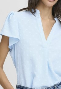 Blusa azzurra chiara con maniche corte a volant, scollatura a V, tessuto texturizzato con piccole punteggiature in rilievo e una vestibilità ampia.