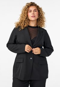 Black blazer with a tailored design, featuring two front pockets, a single button closure, and long sleeves layered over a sheer polka dot top.