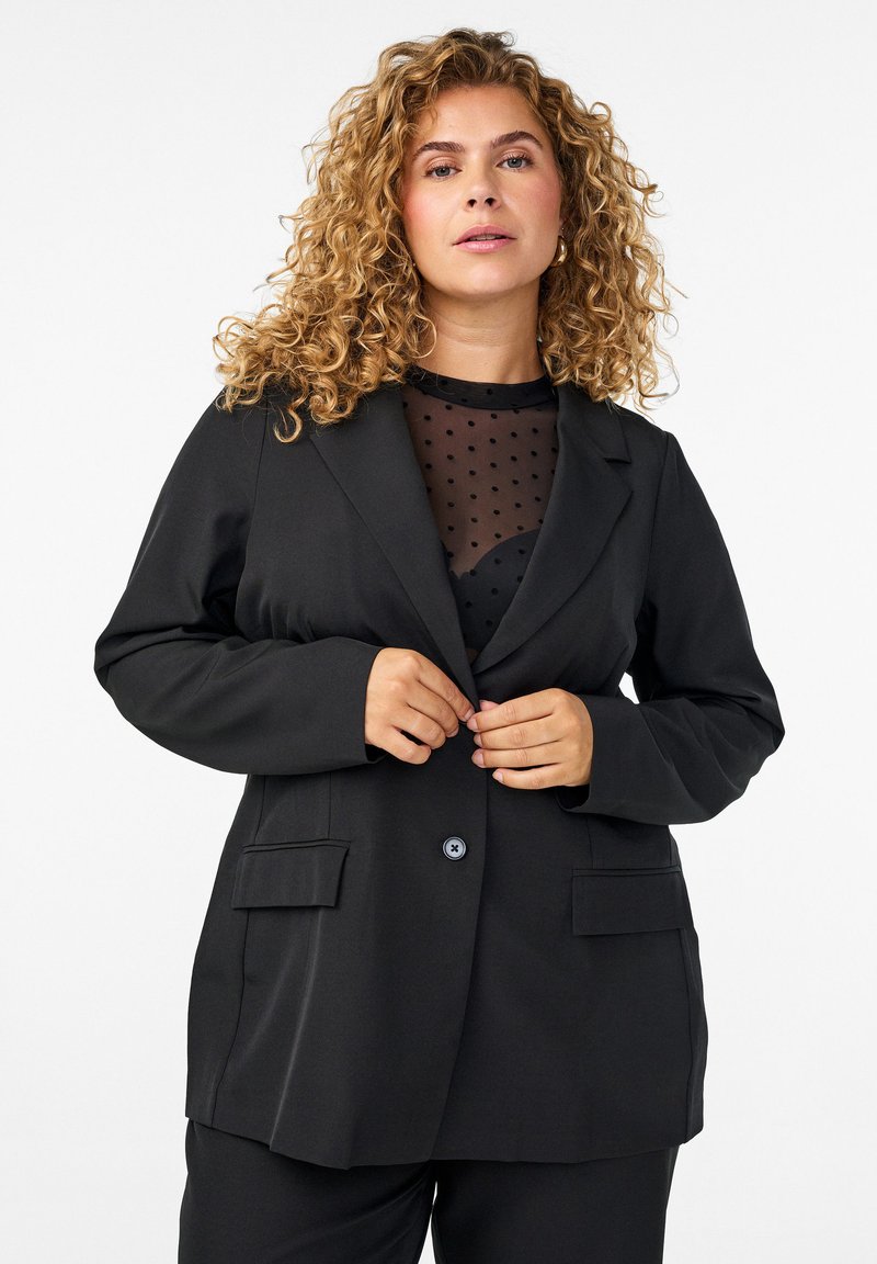 Black blazer with a tailored design, featuring two front pockets, a single button closure, and long sleeves layered over a sheer polka dot top.