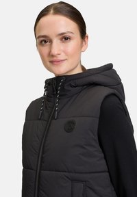 Schwarze wattierte Weste mit Kapuze, ausgestattet mit einem Frontreißverschluss, verstellbaren Kordeln und einem runden Logopatch. Glatte Textur und warme Isolierung.