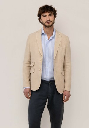 Hombre con cabello corto y rizado, vestido con blazer beige, camisa azul claro con rayas y pantalones oscuros, de pie frente a un fondo liso.