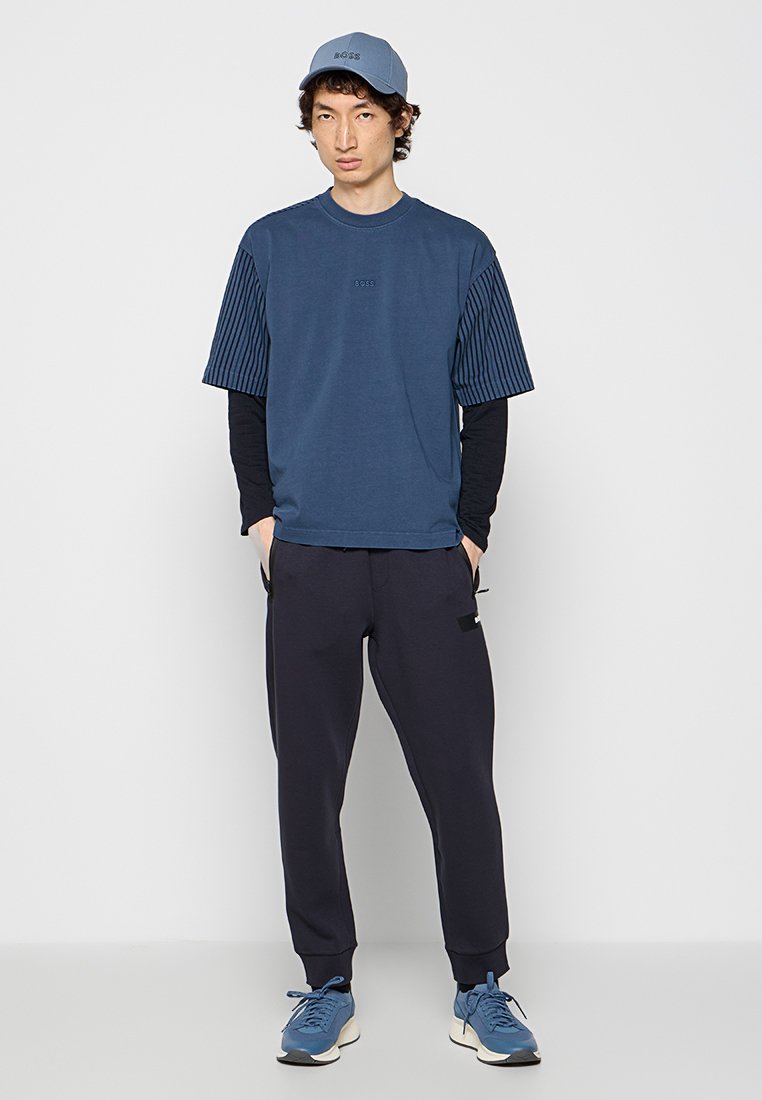 Marine T-shirt met gestreepte mouwen, gecombineerd met donkere pants. Het model draagt een bijpassende lichtblauwe pet en blauwe sport schoenen.