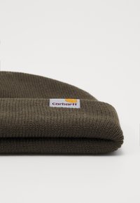Beanie in maglione khaki con risvolto e un'etichetta rettangolare tessuta con il logo Carhartt in arancione e mogano. Texture morbida.