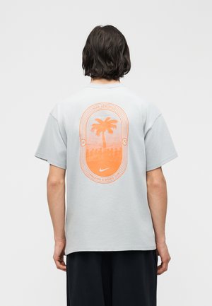 Lühikeste tumedate juustega inimene, kes kannab heledat halli Nike t-särki oranži palmipuud kujutava graafika ja tekstiga seljal.