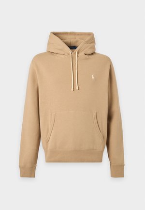 Jeune homme portant un sweat à capuche beige, un pantalon gris foncé et des baskets blanches, debout devant un fond blanc uni.
