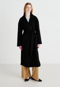 Μαύρο παλτό με ζώνη, πλατιές πέτες και πλαϊνές τσέπες, τοποθετημένο πάνω από πλισέ χρυσά παντελόνια, συνδυασμένο με μαύρα φλατ παπούτσια.