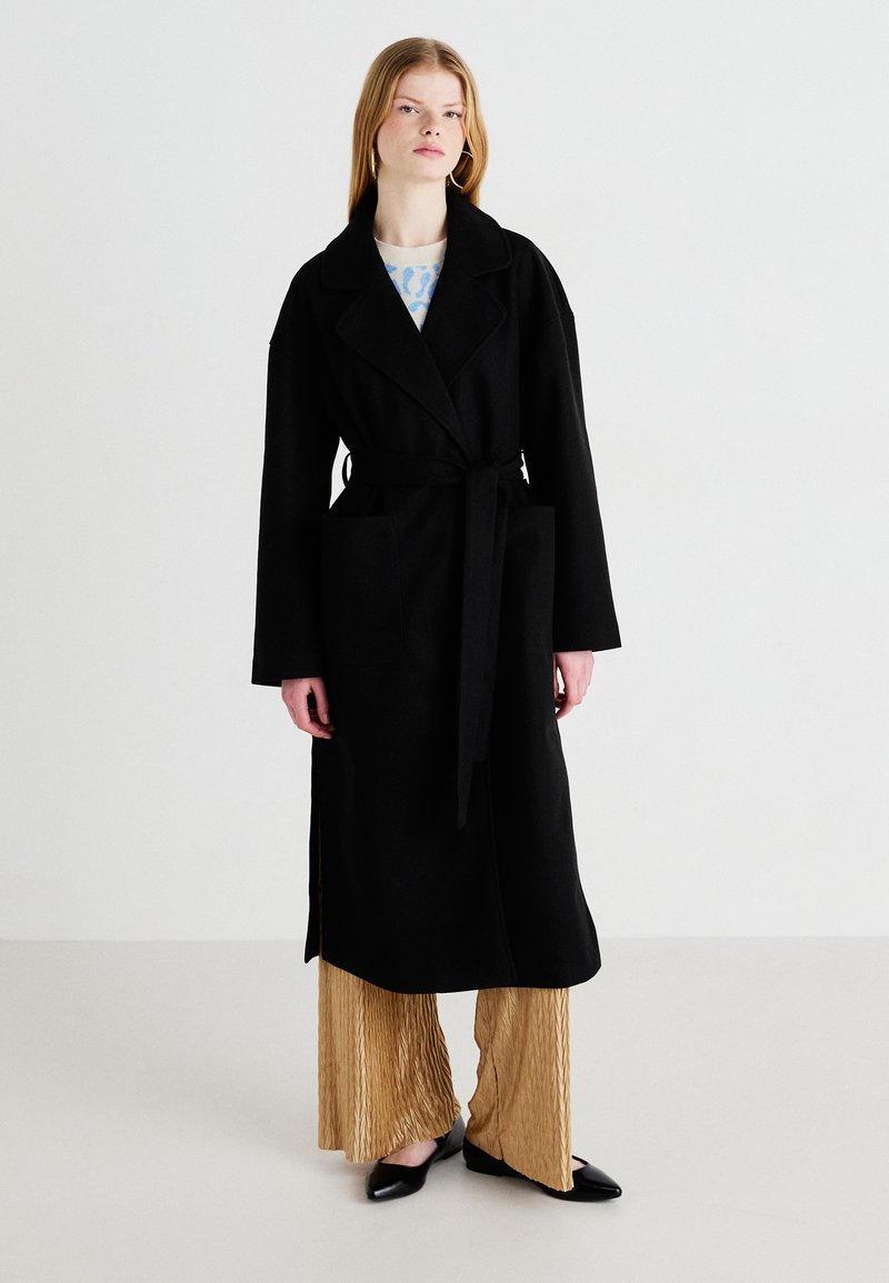 Μαύρο παλτό με ζώνη, πλατιές πέτες και πλαϊνές τσέπες, τοποθετημένο πάνω από πλισέ χρυσά παντελόνια, συνδυασμένο με μαύρα φλατ παπούτσια.