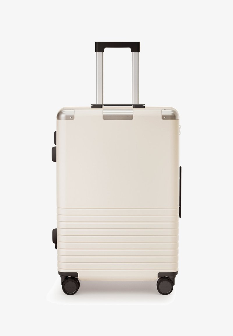 Harde koffer in crème, met een telescopische hendel, zwarte wielen, metalen accenten en horizontale ribbels op het oppervlak.