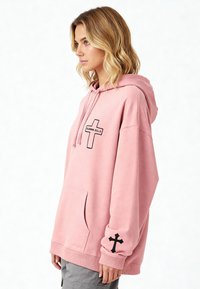 Felpa rosa realizzata in tessuto morbido, con un design a croce nera sul petto e sui polsini. Include una tasca a marsupio e un cappuccio regolabile.